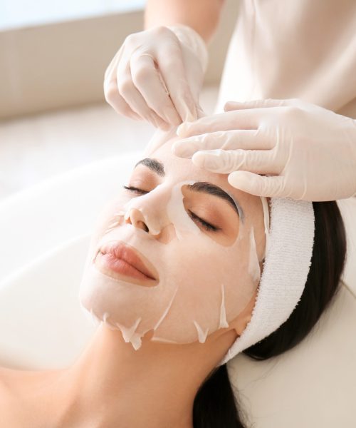 Cosmetologist applying sheet mask on woman's face in beauty salon